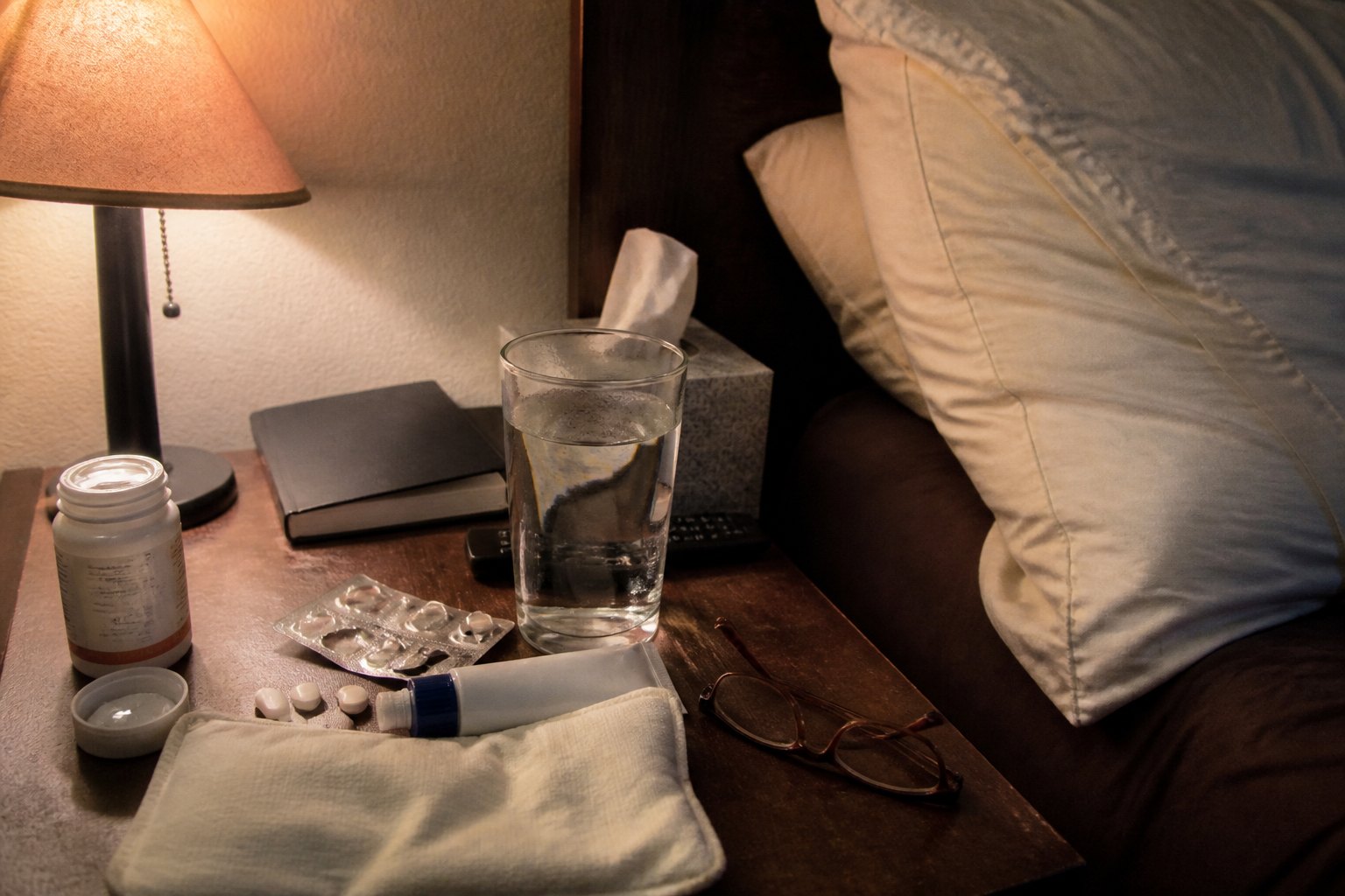 A nightstand covered in painkillers, rubs, heat packs and an old pillow